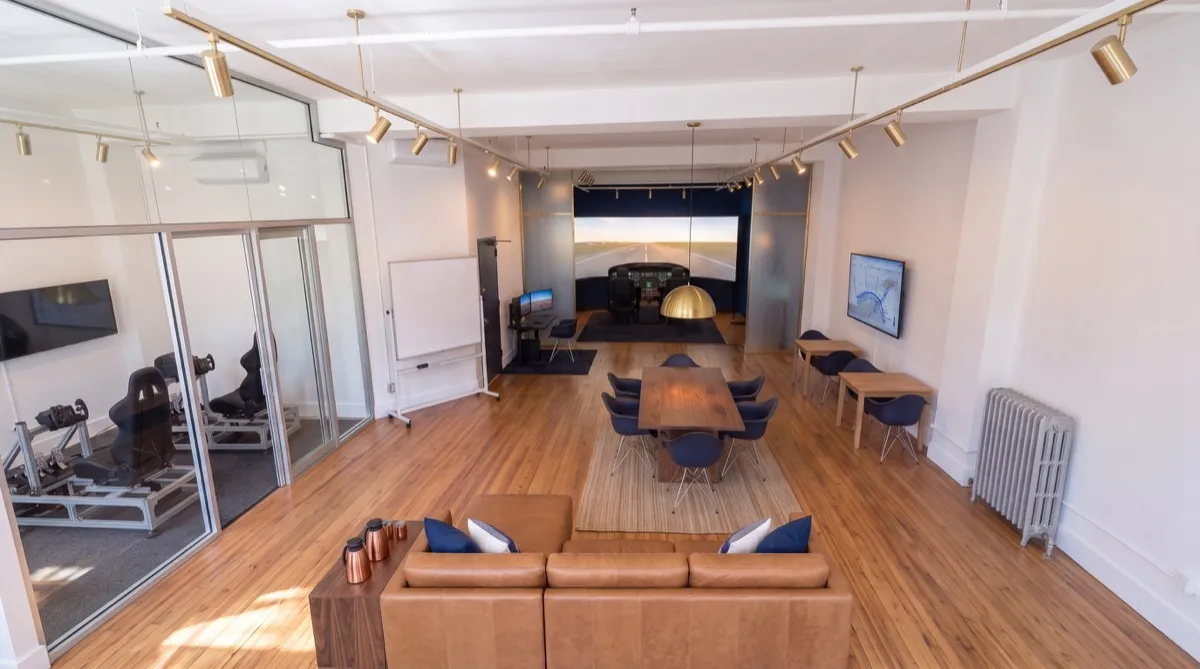 AI-generated rendering of the planned Aviator.NYC training studio in Manhattan seen from above: conference and lounge area by the street windows, two VR motion simulators in the glass room, briefing tables, and the Cirrus G3000 simulator bay at the back