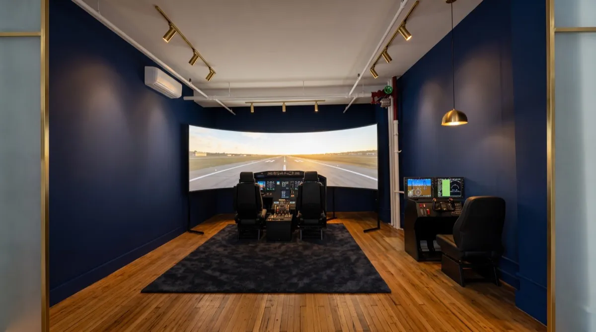 AI-generated rendering of the Cirrus G3000 simulator bay at the Aviator.NYC studio: a 10-foot wrap-around visual showing a runway, dual cockpit seats and console, deep navy walls, and warm accent lighting