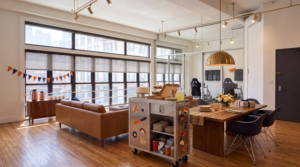 AI-generated rendering of the window-end lounge of the planned Midtown studio set up for a party, with cake, cupcakes and pizza on the sticker-covered airline galley cart beside the conference table