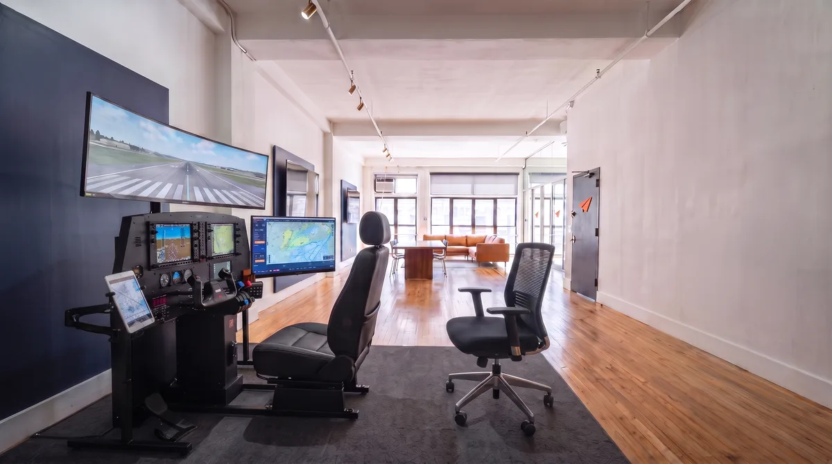AI-generated rendering of the G1000 flight simulator station and its curved ultrawide visual display in the planned Midtown studio, looking down the loft toward the window-end lounge with the glass-walled VR room at the far end